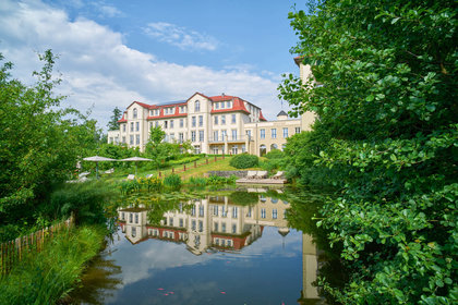 Ein elegantes Gebäude spiegelt sich in einem ruhigen Teich, umgeben von grüner Natur und Bäumen.