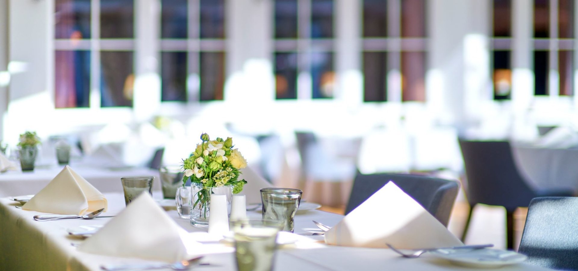 Tables are elegantly laid with floral decorations in a light-flooded room. 