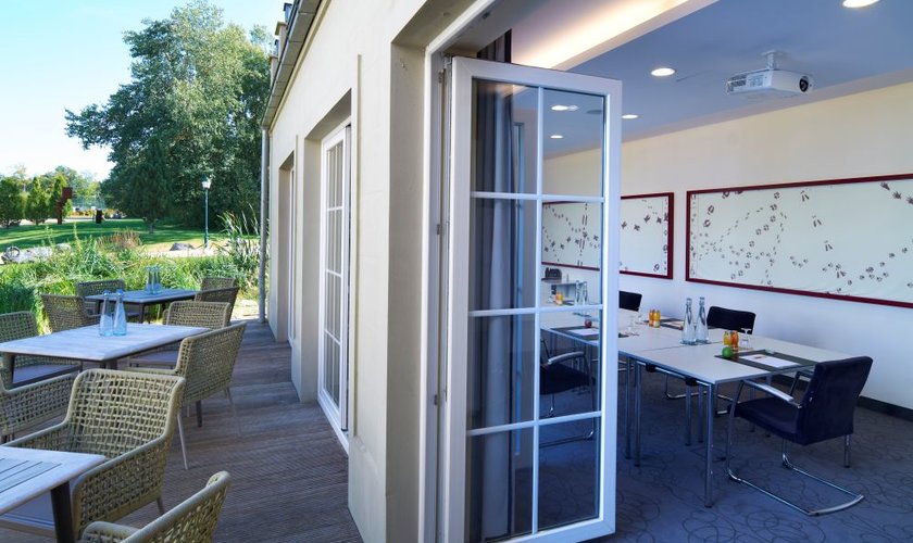 Meeting rooms close to nature Ein Tagungsraum von außen fotografiert, bei dem die Bodenlangen Türen und Fenster offen stehen und im Hintergrund die grünen Bäume stehen.