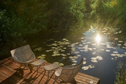 Zwei Rattansessel mit Beistelltischen stehen auf einem Holzsteg am Teich